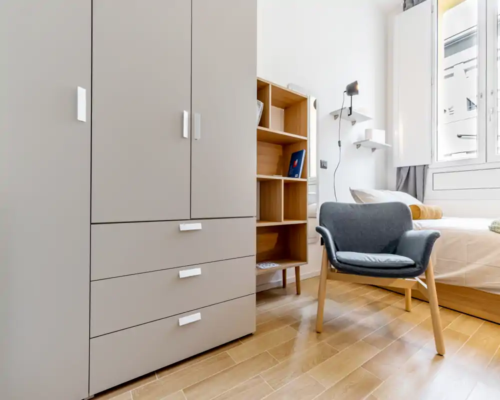 Coin de chambre lumineux et moderne avec lit visible, fauteuil confortable, étagères et grand placard; sol en bois chaleureux et lumière naturelle créent une ambiance accueillante.