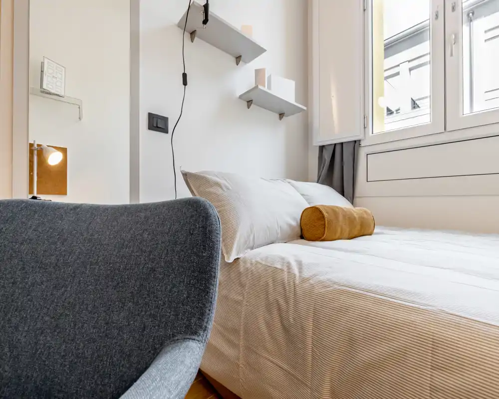 Chambre lumineuse et moderne avec un lit bien fait, coussin décoratif et lumière naturelle provenant d'une grande fenêtre — élégante et accueillante.