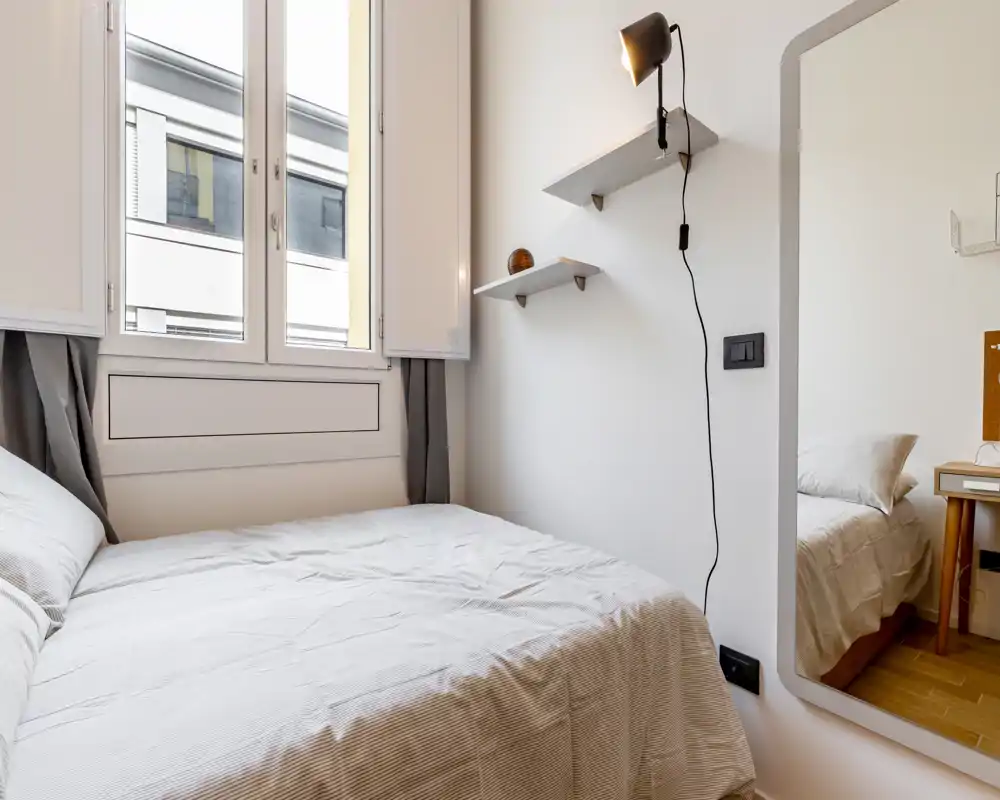 Camera da letto luminosa e minimal con letto matrimoniale ben rifatto sotto una grande finestra, mensole semplici a parete e uno specchio che riflette la luce — perfetta per un affitto moderno e accogliente.
