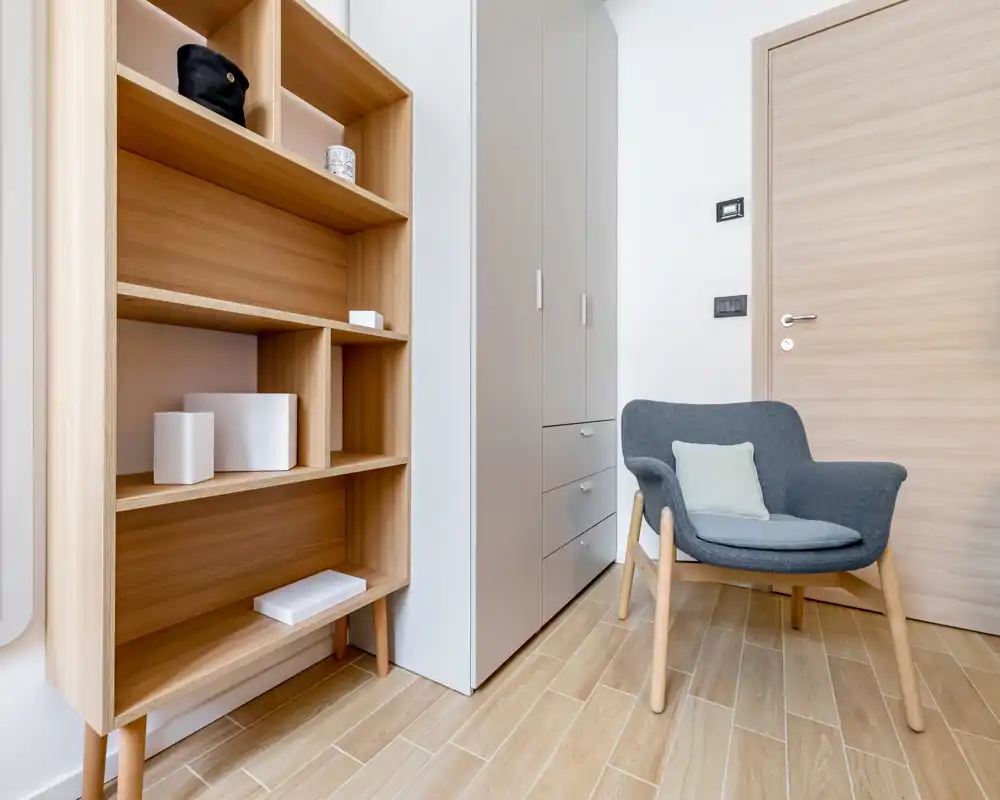 Angolo seduta moderno e ordinato con poltrona grigia, scaffalatura in legno e guardaroba integrato — luminoso e minimalista.