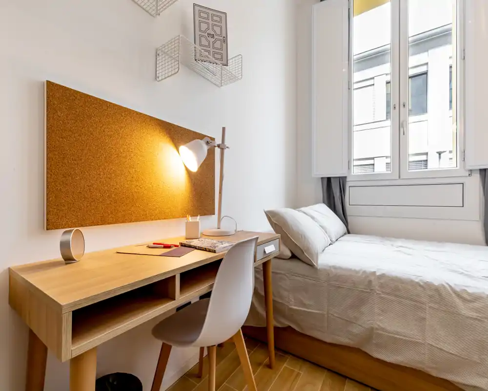 Camera compatta e luminosa con letto singolo vicino a una grande finestra e una scrivania in legno con lampada da lavoro — ideale per studenti o soggiorni brevi.