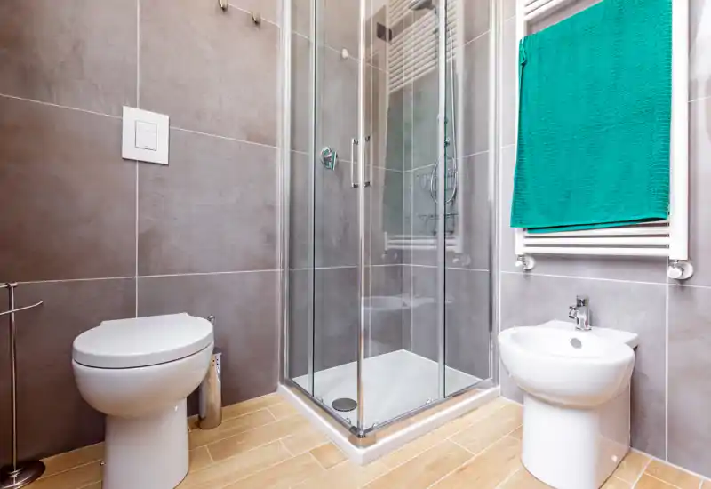 Modern, compact bathroom with a glass shower enclosure, toilet and bidet, light wood-effect floor and grey tiled walls; bright and well-maintained.