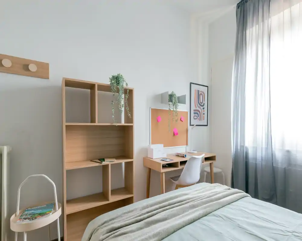 Dormitorio luminoso y minimalista con una cama cómoda en primer plano, un escritorio de madera con corcho y una estantería; la luz natural filtrada por las cortinas aporta un rincón tranquilo para estudiar y descansar.