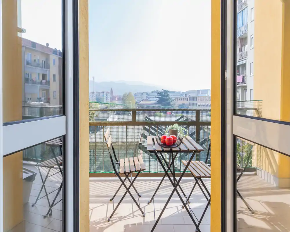 Balcón privado soleado con una pequeña mesa bistró de madera y dos sillas plegables, con vistas a edificios vecinos y colinas lejanas — ideal para el café de la mañana.