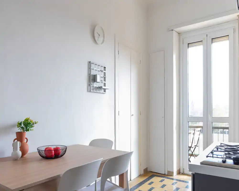 Área de comedor luminosa y minimalista con mesa de madera, cuatro sillas blancas, jarrón decorativo y cuenco de fruta, y grandes puertas de cristal que comunican con el balcón aportando mucha luz natural.