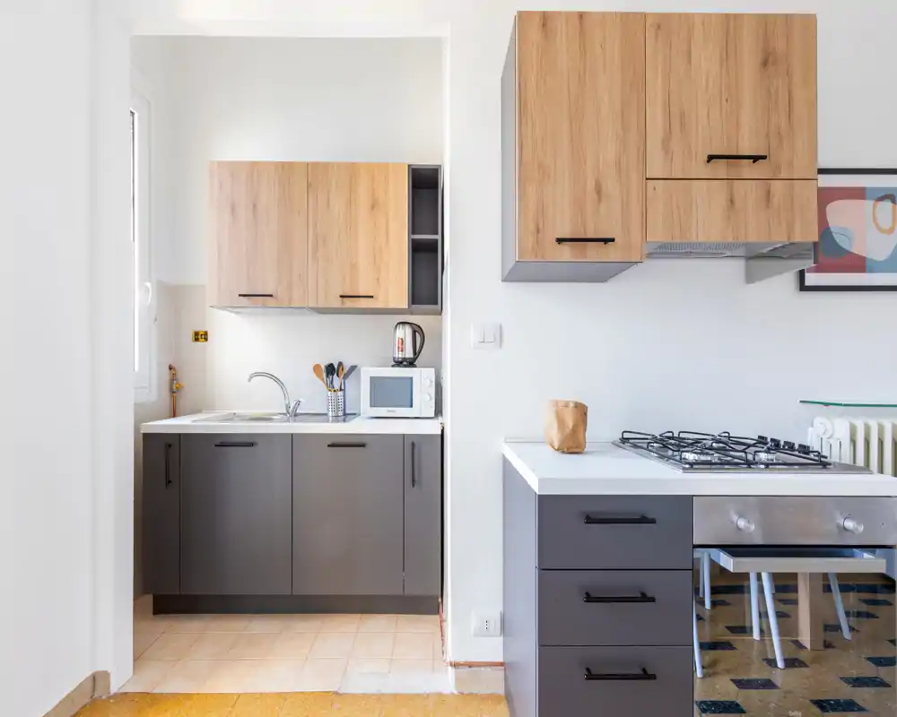 Cocina moderna y compacta con muebles bajos grises y armarios altos de madera, placa de gas y pequeño fregadero con microondas — luminosa, limpia y bien presentada para anuncios de alquiler.