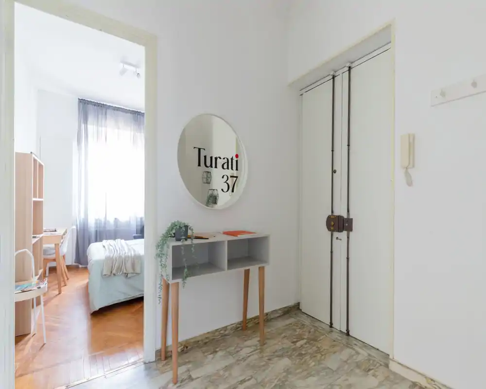 Entrada luminosa y minimalista que se abre a un dormitorio con la cama bien hecha y un pequeño escritorio junto a la ventana. Paredes claras, suelo tipo mármol y un espejo redondo dan una sensación limpia y aireada.