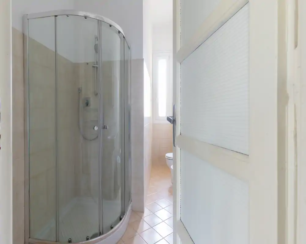 Baño limpio y luminoso con ducha de vidrio curva y suelo de baldosas; luz natural procedente de una ventana estrecha junto al inodoro.