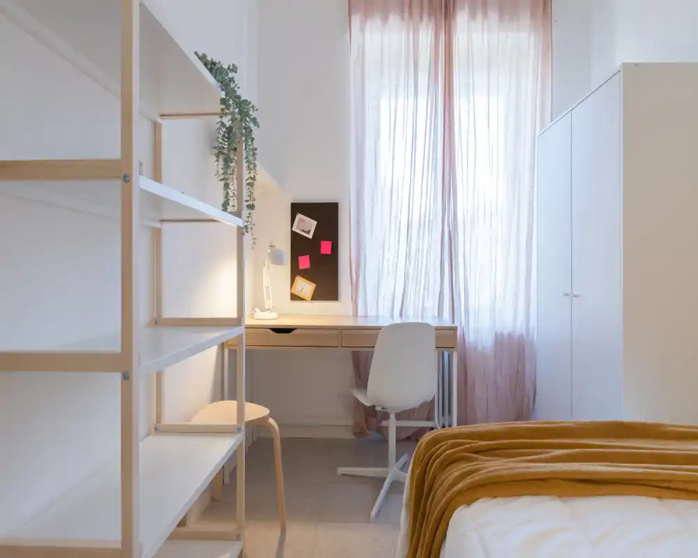 Chambre lumineuse et minimaliste avec un plaid moutarde sur le lit, un bureau bien rangé sous des voilages et une armoire — idéale pour travailler et se détendre.