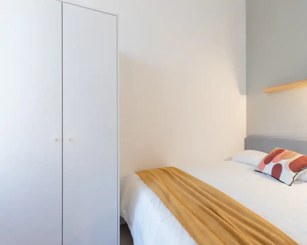 Chambre lumineuse et minimaliste avec un lit bien fait, un coussin décoratif et un plaid moutarde ; murs blancs et armoire simple pour une ambiance moderne et apaisante.