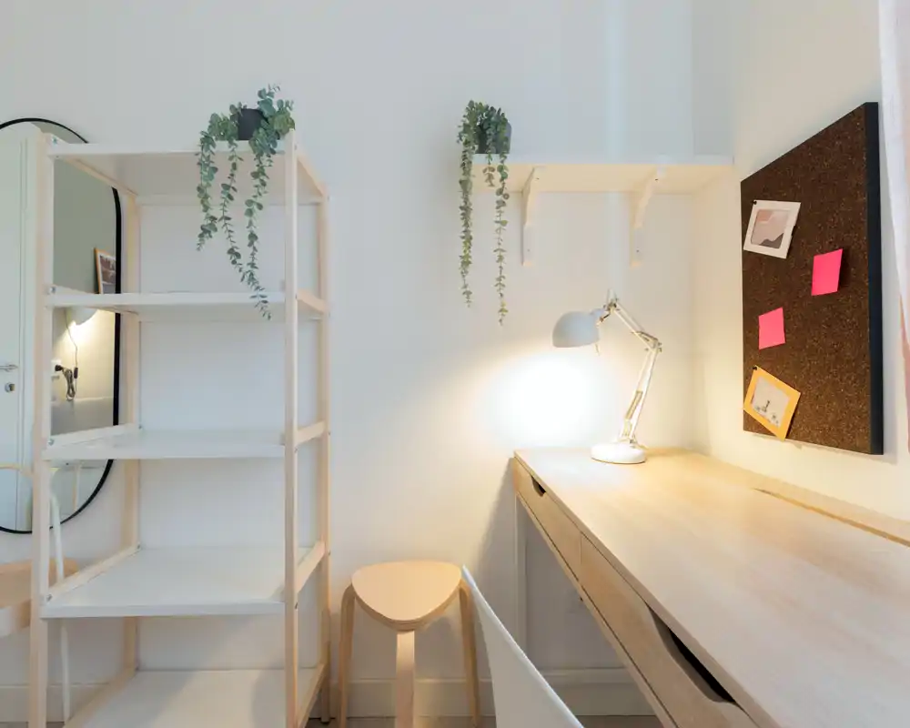 Espace de travail lumineux et minimaliste avec un bureau en bois clair, lampe de bureau, tableau en liège et étagères ouvertes agrémentées de plantes suspendues — parfait pour le travail ou les études.