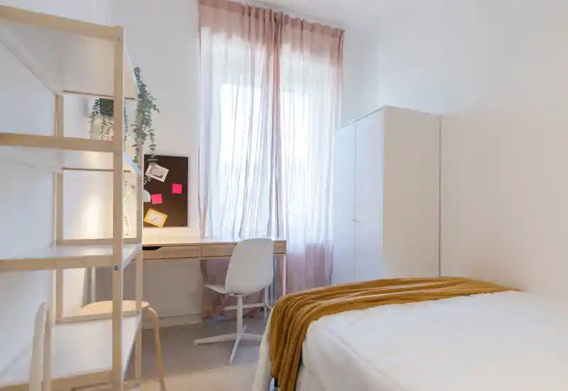 Chambre lumineuse et épurée avec lit blanc, plaid moutarde et un petit bureau près d'une grande fenêtre aux voilages légers.