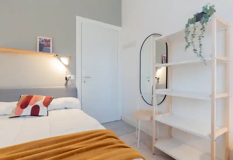 Chambre moderne et lumineuse avec lit bien fait, coussin décoratif et plaid chaleureux ; étagères en bois clair et miroir ovale pour un style minimaliste.