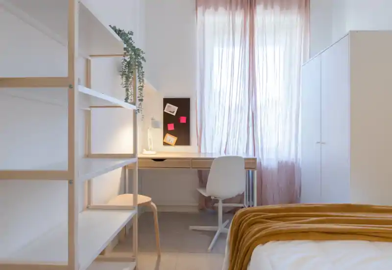 Chambre lumineuse et minimaliste avec un plaid moutarde sur le lit, un bureau bien rangé sous des voilages et une armoire — idéale pour travailler et se détendre.