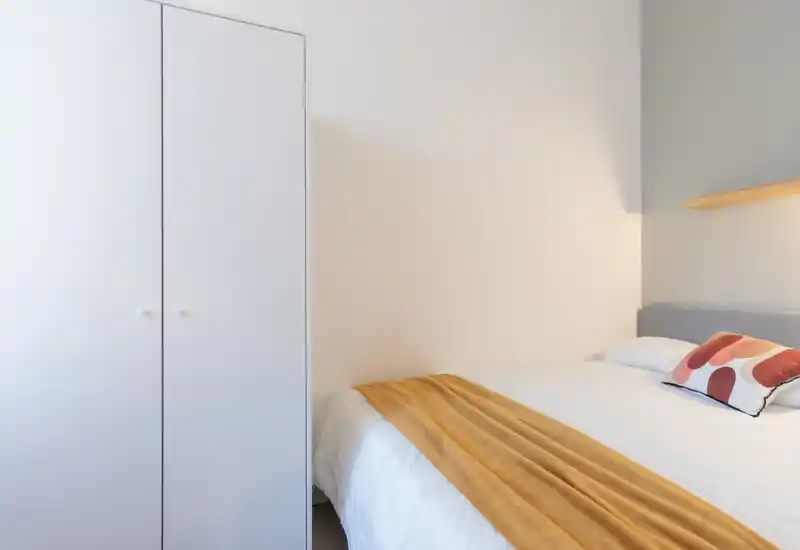 Chambre lumineuse et minimaliste avec un lit bien fait, un coussin décoratif et un plaid moutarde ; murs blancs et armoire simple pour une ambiance moderne et apaisante.
