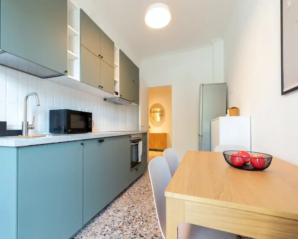 Cuisine moderne en enfilade avec meubles bas vert, four et micro-ondes intégrés, table en bois et crédence carrelée lumineuse — espace propre et fonctionnel parfait pour les locations courte durée.