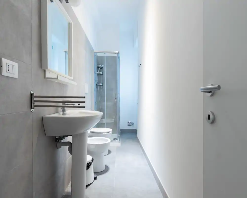 Salle de bains moderne et lumineuse avec lavabo sur pied, WC, bidet et douche vitrée. Carrelage gris et lignes épurées pour un style minimaliste et frais.