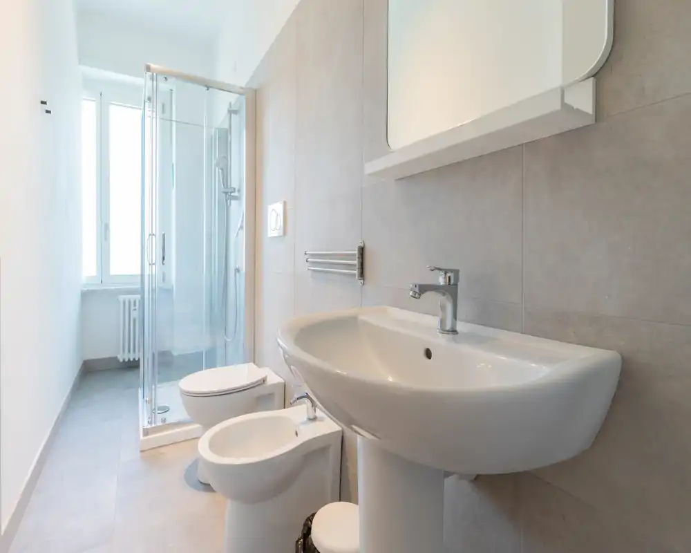 Salle de bains moderne et lumineuse avec lavabo sur pied, bidet, WC et douche vitrée près de la fenêtre. Carrelage neutre et lignes épurées pour une ambiance fraîche et minimaliste.
