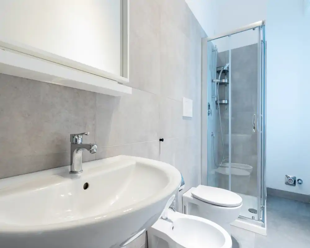 Salle de bains moderne et lumineuse avec lavabo, bidet, WC et douche à l'italienne avec paroi en verre ; carrelage neutre et équipements propres pour un rendu minimaliste.