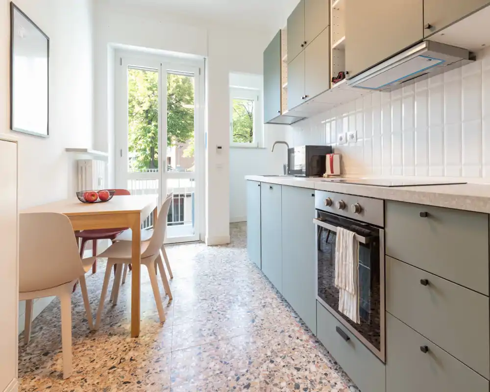 Cuisine moderne et lumineuse avec meubles verts doux, four encastré et petite table près d'une porte vitrée laissant entrer la lumière — un espace de cuisson pratique et accueillant.