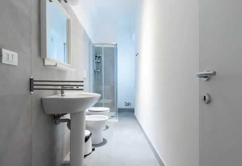 Bright, modern bathroom with a pedestal sink, toilet, bidet and a glass-enclosed shower. Clean lines and neutral grey tiles create a fresh, minimalist look.