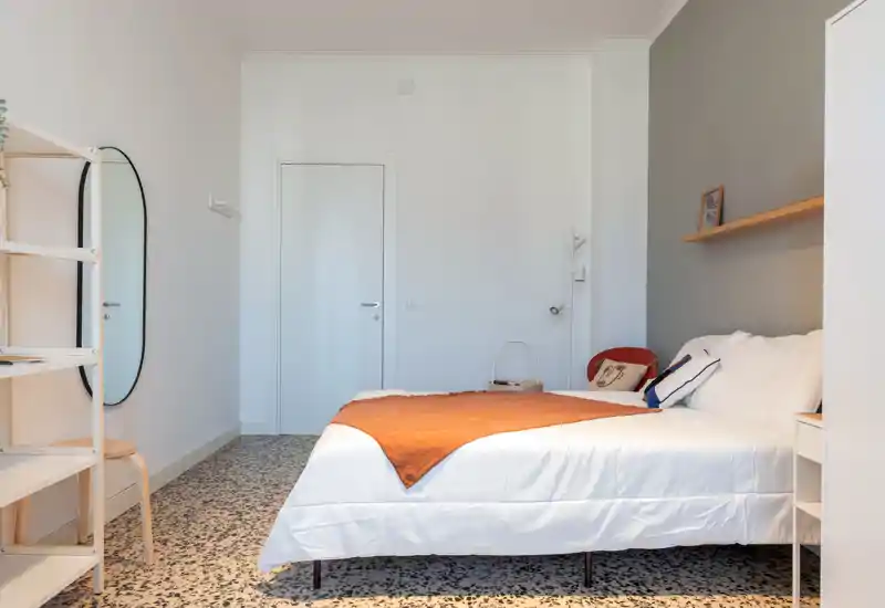 Camera da letto moderna e luminosa con un letto ben rifatto, coperta arancione calda e mensole essenziali. Linee pulite, pavimento in graniglia e luce naturale morbida creano un ambiente accogliente e minimal.