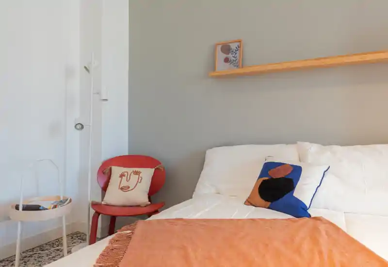 Camera da letto luminosa e accogliente con letto ben rifatto, cuscini decorativi e tessuti caldi. Una mensola semplice e una poltroncina accent pianificano un’immagine invitante per l’annuncio.
