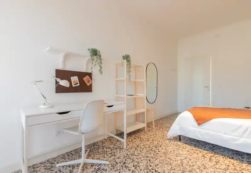 Camera da letto luminosa e minimal con scrivania bianca e scaffale, letto ben rifatto visibile a destra e un caldo plaid arancione. Linee pulite, pavimento in graniglia e piante decorative per un ambiente moderno e rilassante.