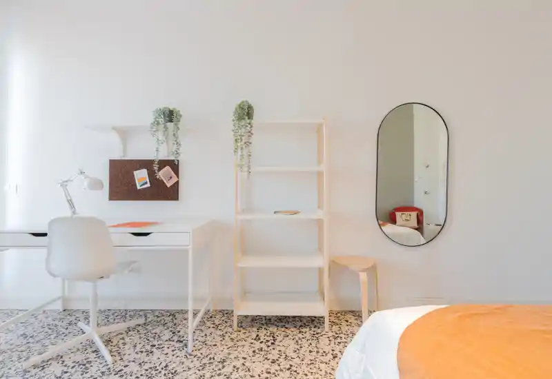 Angolo camera luminoso e minimale con scrivania bianca, scaffalatura aperta e letto con coperta arancione calda. Stile pulito d'ispirazione scandinava e luce naturale rendono l'ambiente accogliente.