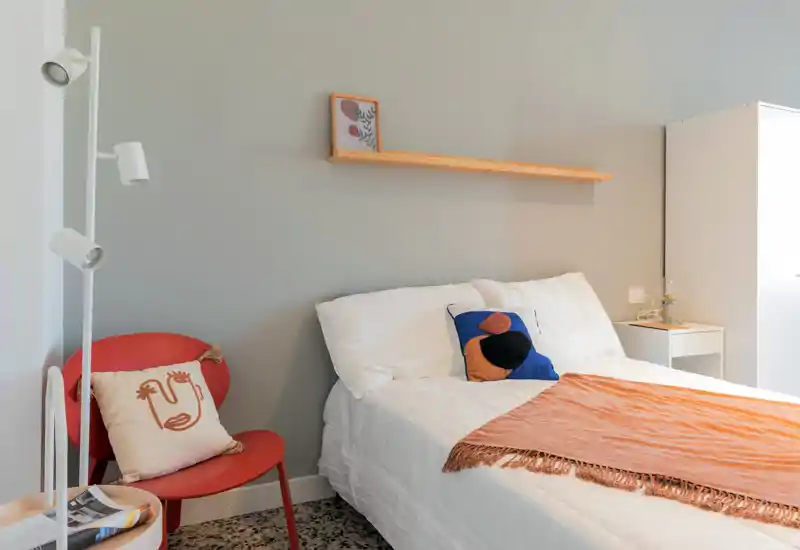 Camera da letto moderna e luminosa con letto ben rifatto, cuscini decorativi e un caldo plaid arancione; mensola minimal e una poltrona per la lettura completano l'ambiente.