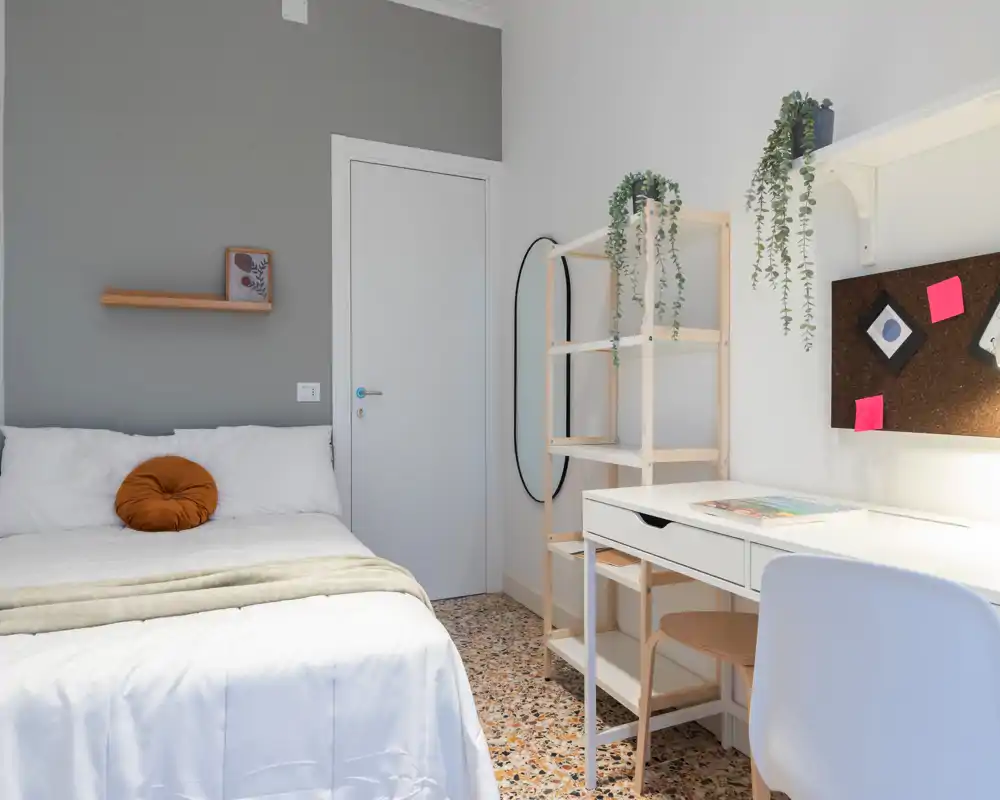 Bright, modern bedroom with a neatly made double bed, decorative round cushion, shelving and a functional white desk — ideal for students or short-term stays.