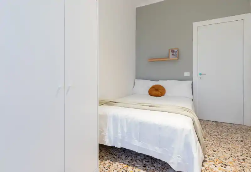 Bright, well-styled small bedroom with a neatly made double bed, neutral tones and a decorative round cushion. Clean minimal design, terrazzo floor and a compact wardrobe maximize space.