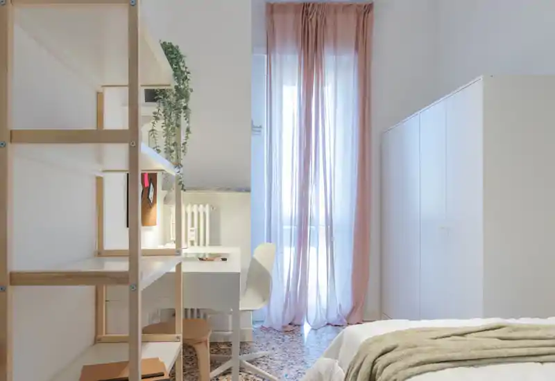 Bright, minimal bedroom with a visible bed in the foreground, a desk and shelving to the left, and soft pink curtains over a balcony door that bring natural light.