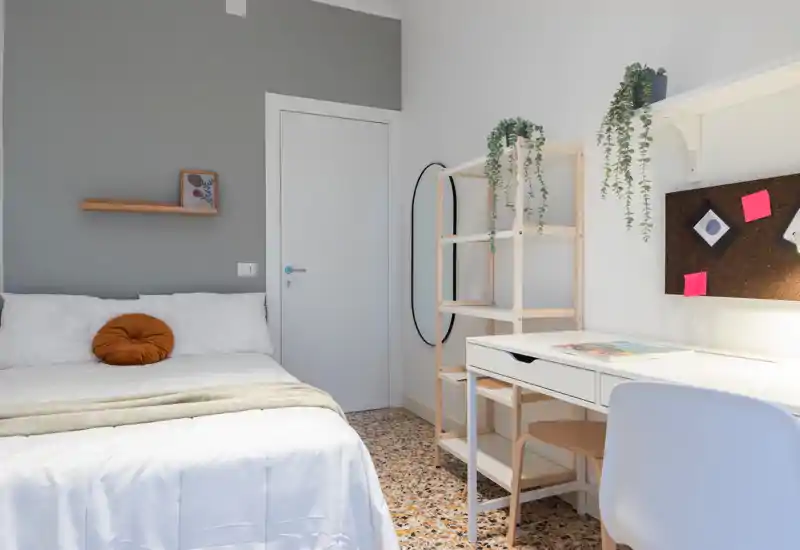 Bright, modern bedroom with a neatly made double bed, decorative round cushion, shelving and a functional white desk — ideal for students or short-term stays.