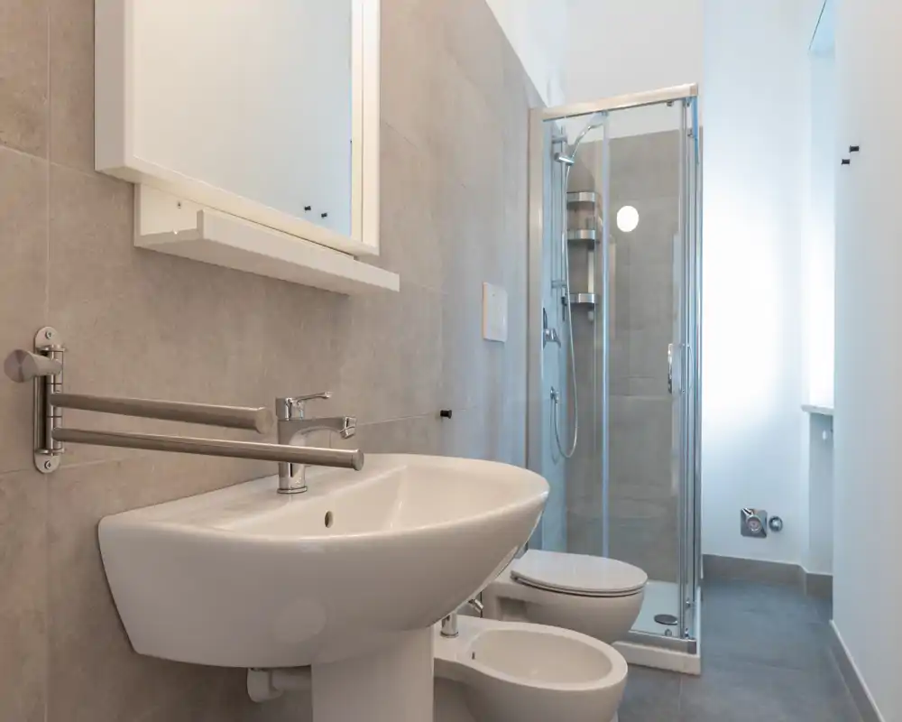 Bagno moderno e luminoso con lavabo a parete, bidet, WC e doccia con box in vetro; piastrelle grigie neutre e arredi puliti conferiscono un aspetto fresco e minimale.