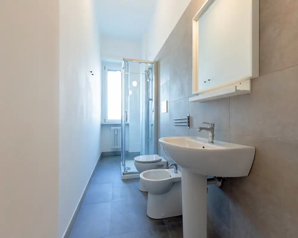 Bagno moderno e compatto con lavabo a colonna, bidet, wc e box doccia angolare in vetro; piastrelle grigie neutre e specchio retroilluminato per un aspetto pulito e luminoso.
