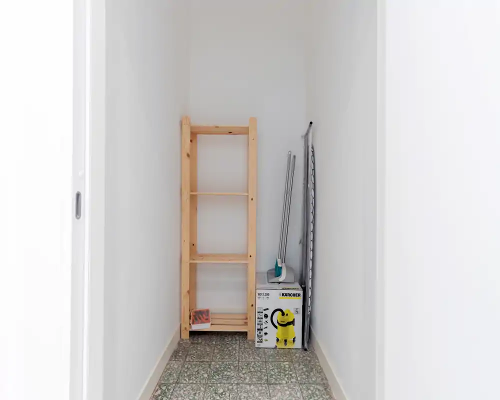 Piccolo ripostiglio stretto con un semplice scaffale in legno e attrezzatura per le pulizie; adatto a essere descritto come un armadietto per utensili o un angolo deposito.