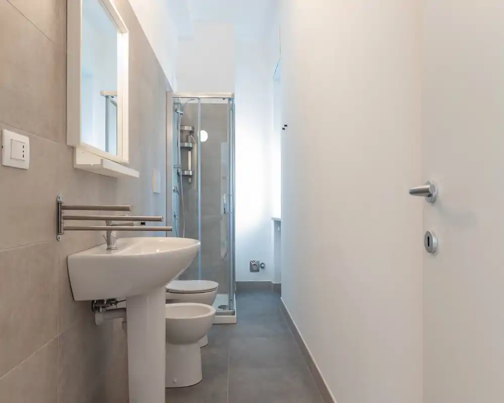 Bagno moderno e compatto con lavabo a colonna, WC, bidet e doccia con box in vetro. Piastrelle grigie neutre e luce naturale intensa donano un aspetto pulito e contemporaneo.