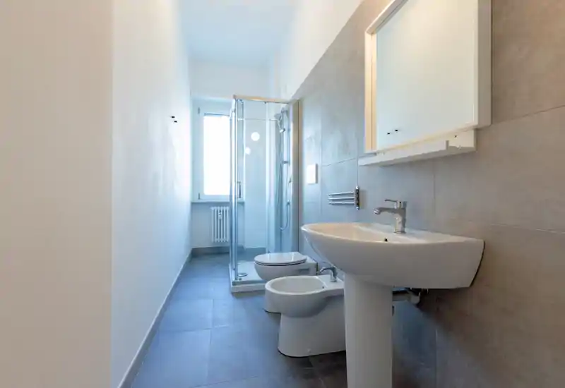 Bright, modern bathroom with a glass shower cubicle, pedestal sink, bidet and toilet, finished in neutral gray tiles and a large mirror.