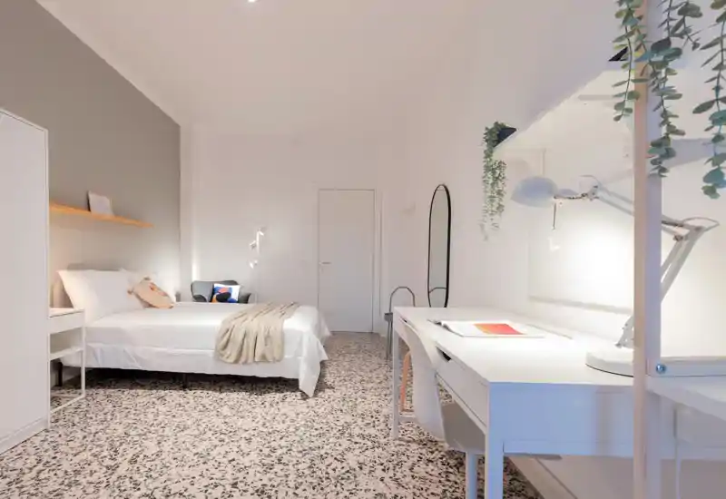 Bright, modern bedroom with a neatly made double bed, bedside table and a well-lit workspace. Minimalist decor, terrazzo-style floor and hanging plants create a fresh, inviting atmosphere ideal for rental photos.
