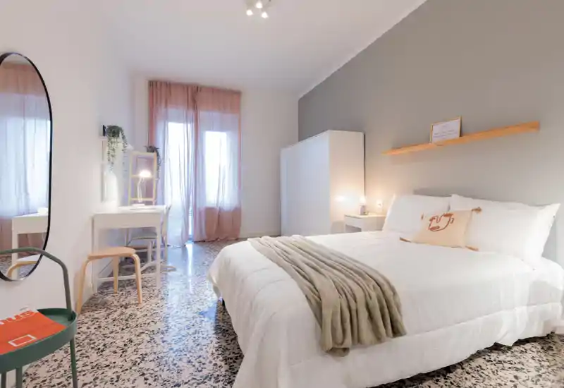 Bright, stylish bedroom with a large white bed, cozy throw and soft natural light from a window with sheer curtains. Clean, modern decor with a dressing desk and wardrobe, ideal for rental photography.