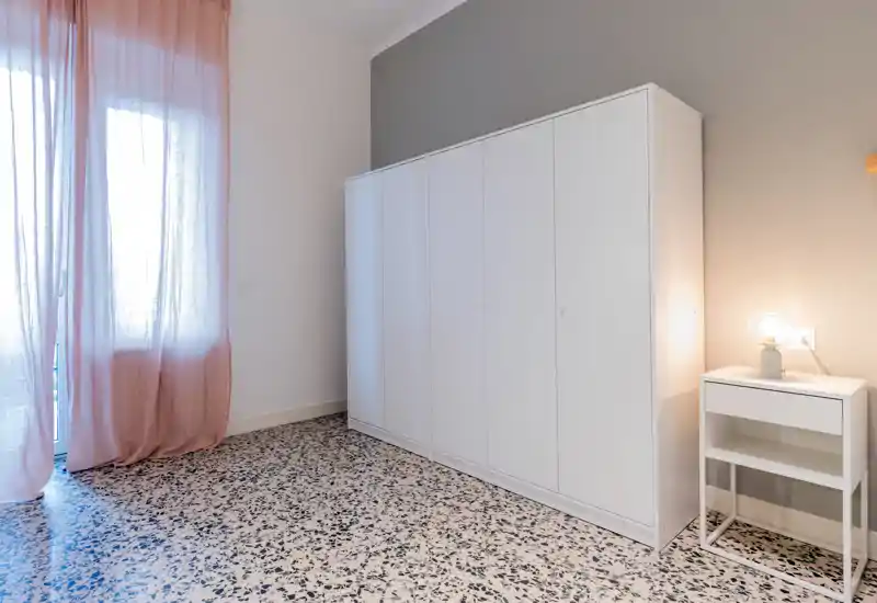 Image shows a corner with a tall white wardrobe, a small bedside table with a lamp and sheer pink curtains; no bed or other defining elements are visible to classify as a bedroom or other specific room.