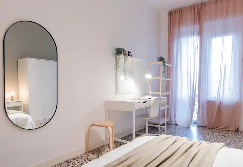 Bright, softly styled bedroom with a visible bed in the foreground, a minimalist desk and a large window with sheer pink curtains letting in natural light.