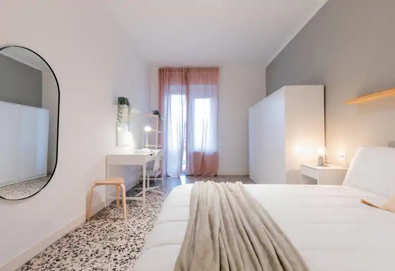 Bright, stylish bedroom with a large bed dressed in neutral linens, soft throw, a bedside lamp and a workspace by the window with sheer pink curtains providing warm natural light.