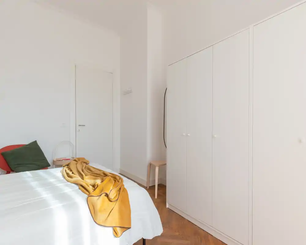 Camera da letto luminosa e ariosa con letto in biancheria bianca, plaid senape e cuscini decorativi; grande armadio bianco e pavimento in legno per un aspetto moderno e tranquillo.