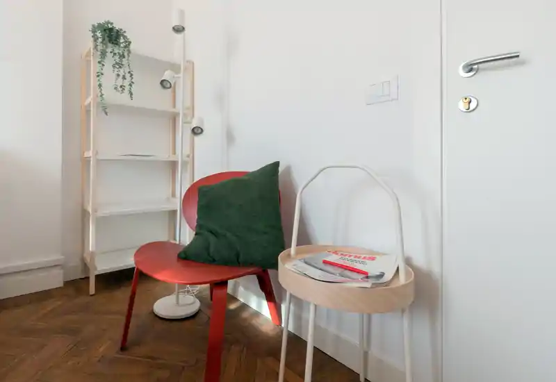 Cozy seating corner with a red accent chair, green cushion, small round side table holding magazines and a light shelving unit with a hanging plant — a compact, well-styled living or entry nook.