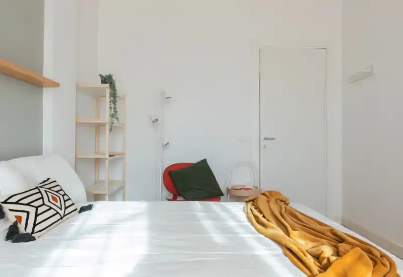 Camera da letto luminosa e minimalista con letto ben fatto, cuscini decorativi e un caldo plaid senape; la luce naturale illumina l'ambiente e valorizza l'arredamento dallo stile nordico.