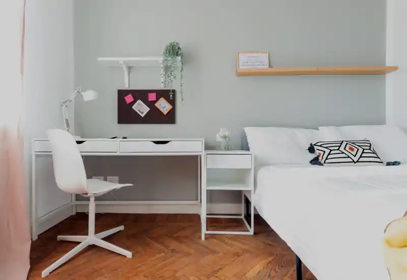 Camera da letto luminosa e minimale con letto bianco e scrivania adiacente in bianco; luce naturale soffusa, cuscino decorativo geometrico e parquet in legno creano un’atmosfera moderna e rilassante.