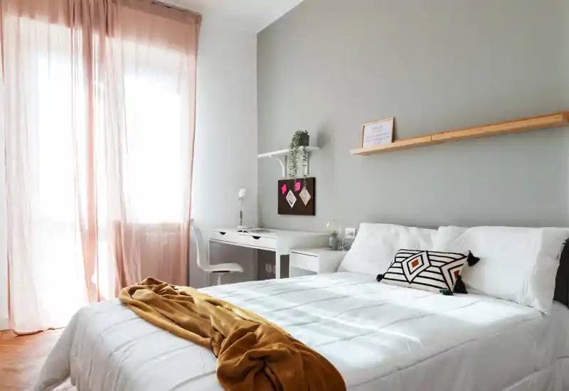 Camera da letto luminosa e di stile con letto matrimoniale ben rifatto, cuscino decorativo e plaid caldo, più una piccola scrivania vicino alla finestra soleggiata — perfetta per soggiorni rilassanti o smart working.