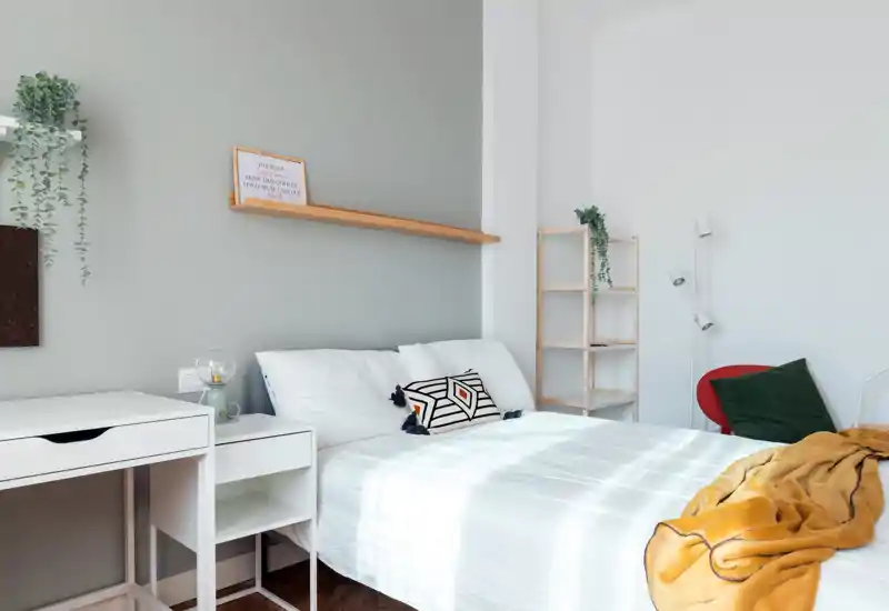 Camera da letto luminosa e moderna con letto matrimoniale rifatto, biancheria bianca, cuscino geometrico e morbida coperta senape; scrivania e scaffali minimal rendono lo spazio pulito e arioso.